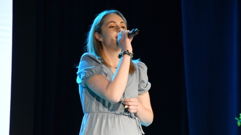 
                                        Pani w blond włosach niebieskiej sukience stoi na scenie z mikrofonem w ręce                                        