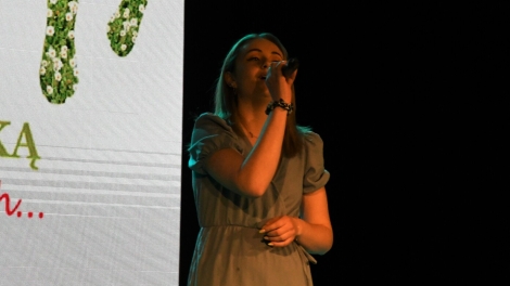 
                                        Na scenie stoi pani w blond włosach zielonej sukience w dłoni trzyma mikrofon                                        