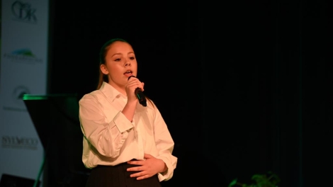 
                                        Na scenie stoi dziewczyna w blond włosach białej koszuli czarnej spódnicy w dłoni trzyma mikrofon                                        