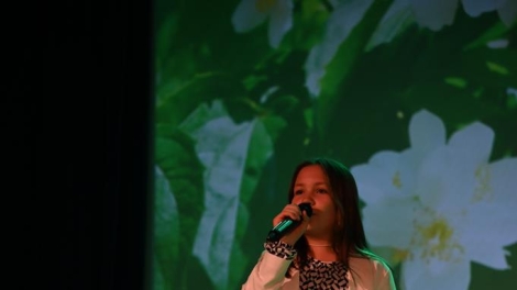 
                                        Na scenie wystepuje dziewczyna w szatynowych włosach beżowej kamizelce biało czarnej sukience w dłoni trzyma mikrofon                                        