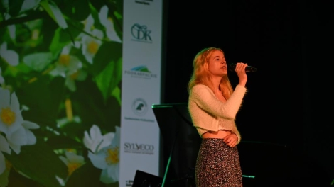 
                                        Na scenie stoi dziewczyna w blond włosach białym swetrze kolorowej spódnicy w dłoni trzyma mikrofon jej usta są otwarte                                        