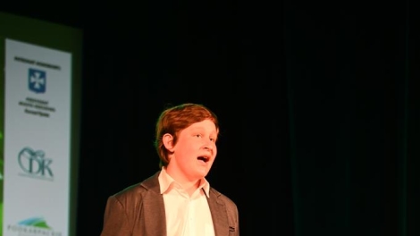 
                                        Na scenie stoi chłopak w szatynowych włosach brązowej marynarce białej koszuli czarnych spodniach jego usta są otwarte                                        