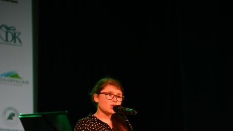 
                                        Dziewczyna w szatynowych włosach okularach czarnej sukience w białe kropki stoi przed mikrofonem                                        