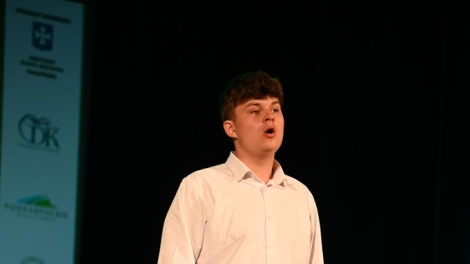 
                                        Na scenie stoi chłopak w szatynowych włosach bialej kozuli czarnych spodniach jego usta są szeroko otwarte                                        