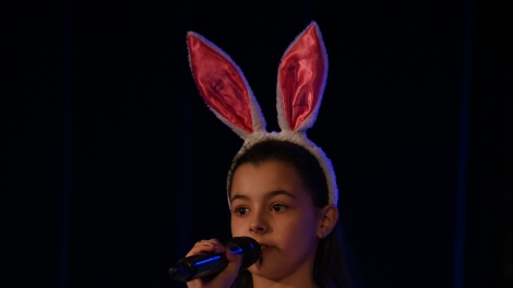 
                                        Na scenie dziewczynka w brunatnych włosach opasce z uszami królika białym swetrze białej spódnicy                                        