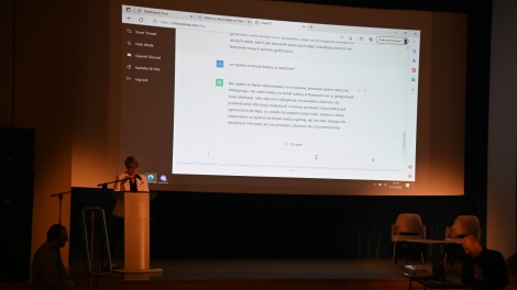 
                                        Na scenie stoi przy mównicy pani w blond włosach białym żakiecie na ścianie wyświetla się przeglądarka internetowa z włączonym programem sztucznej inteligencji                                        