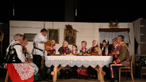 
                                        Przy wiejskim stole siedzą aktorzy pan w białym stroju kroi chleb                                        