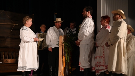 
                                        Osiem osób stoi na scenie ubranych w stroje ludowe dwóch panów z tyłu gra na harmionach                                        