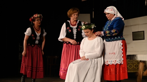 
                                        Na scenie cztery aktorki ubrane w stroje ludowe pani siedzi w białej sukni z wiankiem na głowie                                        