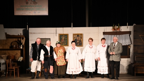 
                                        Na scenie siedem osób ubranych w stroje ludowe z tyłu dekoracja wiejskiej chaty                                        
