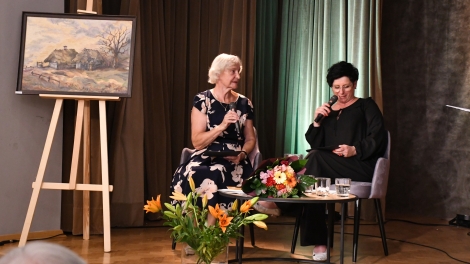 
                                        Na scenie przy stole siedzą Jadwiga Kupiszewska w siwych włosach ciemnej sukni w białe kwiaty obok niej pani w brunatnych włosach czarej sukience w dłoni trzyma mikrofon                                        
