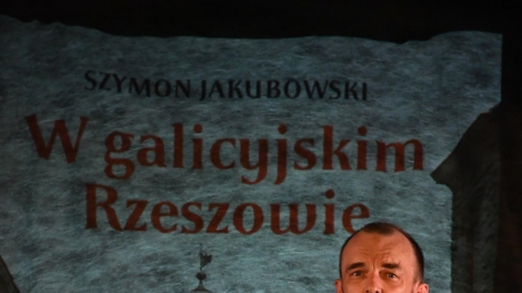 
                                        Szymon Jakubowski stoi na scenie w tle prezentacja jego książki w galicyjskim Rzeszowie                                        