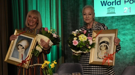 
                                        Dwie panie stoją przy stoliku w rękach trzymają kwiaty i obrazy przedstawiające kobiece twarze                                        