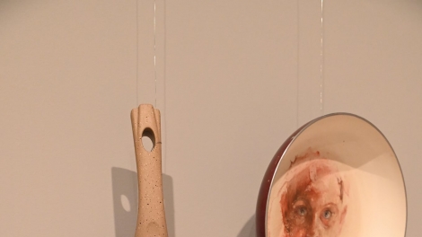 
                                        Na ścianie wystawy wiszą dwie patelnie z namalowanymi na dnie twarzami                                        